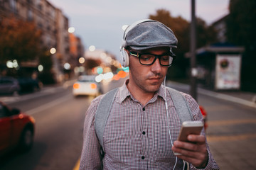 Fashionable retro dressed man with cap, suspenders and eyeglasses standing on city street and using tablet. City lights on sunset.