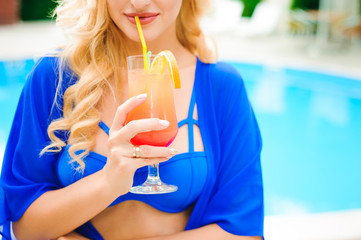 Blonde girl with long hair holding cocktail and posing near pool on the sun.
