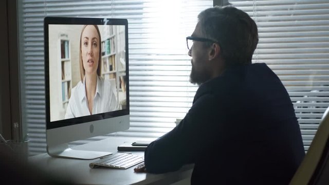 Handheld shot of bearded mature businessman in glasses sitting at office desk and talking to young female colleague via video conversation