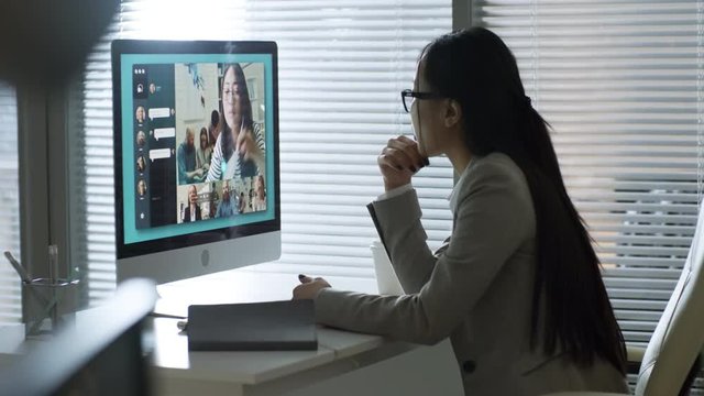 Handheld shot of young Asian woman in glasses talking to group of four colleagues via video conversation in office