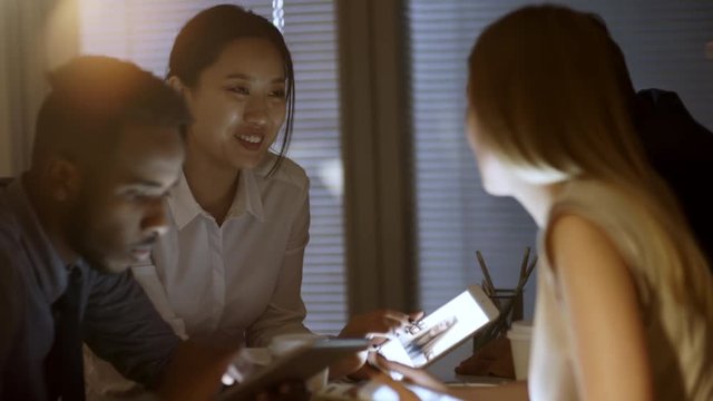 Handheld shot of young Asian woman showing photos on tablet computer to group of multi ethnic friends or colleagues late in the evening