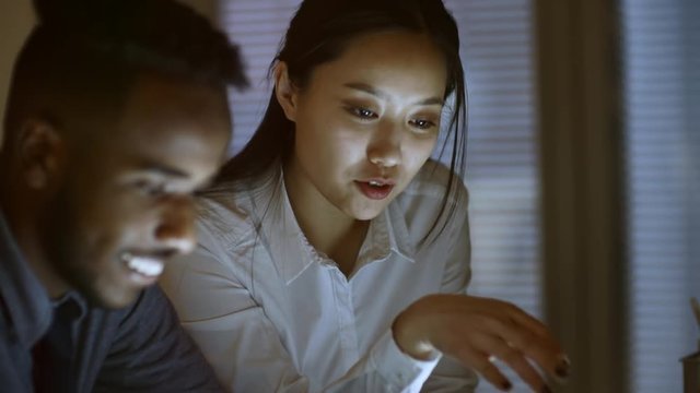Handheld shot of Asian woman talking to black man when using smartphone at office desk late in the evening