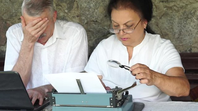senior eldery mature couple at arbor with. man working on laptop and laugh make fun of women who use magnifying glass typing on old vintage typewriter. slow motion static close face outdoor shot