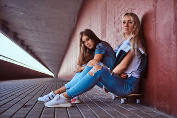 Two beautiful hipster girls sitting on skateboard at a sidewalk under bridge.