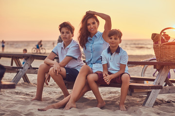 Happy family - mother with her sons sitting on a bench against the background of a seacoast at the bright sunset.