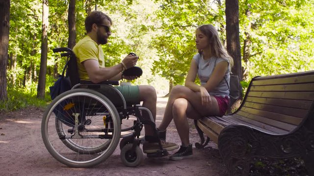 Young disable man in a wheelchair on a walk in the park with his wife. A woman is sitting on a bench. Man smiling