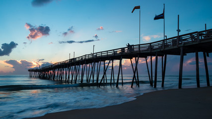 Flags fly in the ocean breeze on an old wooden fishing pier at sunrise. A colorful and dramatic morning sky in the background. Long exposure gives a soft blur to the waves below.