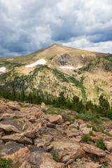 Sunny, largely bare Colorado Rocky Mountains, with dominant pointed peak on left. Patches of snow. Rock field in front.  Slightly dark clouds above.  Good for background, text.