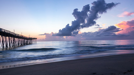 Obraz premium Purple, red and blue sunrise colors light up a calm seashore and tall clouds in the sky. The end of a fishing pier in the distance.