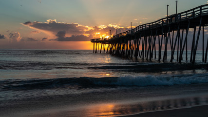 The golden light of a bright sunrise beams through the clouds to light up a fishing pier that extends out into the ocean. Small waves break on the sandy shore. Sunrise colors reflected on the surface.