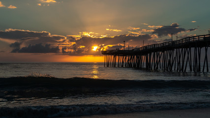 Fototapeta premium A golden sunrise over the Atlantic Ocean in North Carolina. Small waves break on the shore. A fishing pier extends far out into the sea.