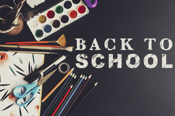 time back to school concept text white chalk on black board, colorful pencils paints brushes scissors and notebook, space for text, flat lay