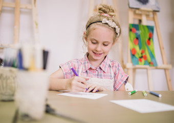 Blonde cute smiling seven-year-old girl, draws at the table in the creative studio