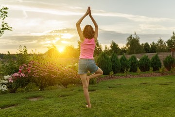 Young girl practicing yoga, meditating on a summer sunset background, on green grass, lawn near the house.