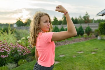 People, power, stamina, strength, health, sport, fitness concept. Outdoor portrait smiling teenage girl flexing her muscles, background green lawn, sunset