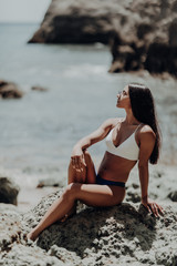 Fototapeta na wymiar Portrait of young beautiful sexy tanned girl in white bikini sitting at rocks near blue green clear ocean sea water. Sunbathing woman.