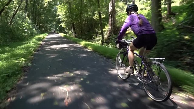 Low angle close behind a mature woman on a mountain bike, cycling on a paved rail trail in a forest.