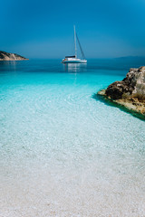 Fototapeta premium Azure blue lagoon with sailing catamaran yacht boat at anchor. Pure white pebble beach, some rocks in the sea