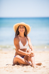 Young latin attractive in white bikini and straw hat posing while sitting on the beach