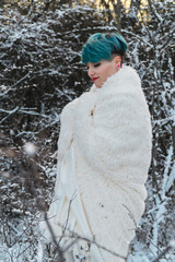 Lovely girl with blue hair in winter wrapped in a white fur coat in a snowy park walks and smiles