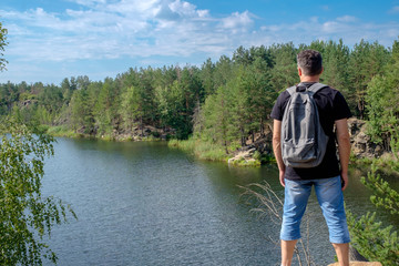 Adult white tourist with backpack stands on the rock and looks at the lake.