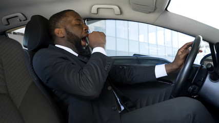 Tired black man yawning in car, overworked businessman driving car, danger