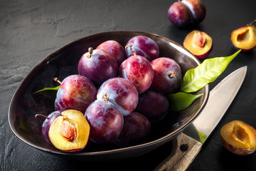 Whole fresh red violet blue plums and slices cut with knife in bowl on kitchen table