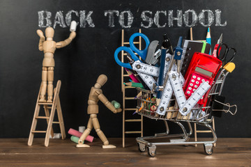Shopping cart / trolley with school supplies and wooden puppets on blackboard background. Back to...