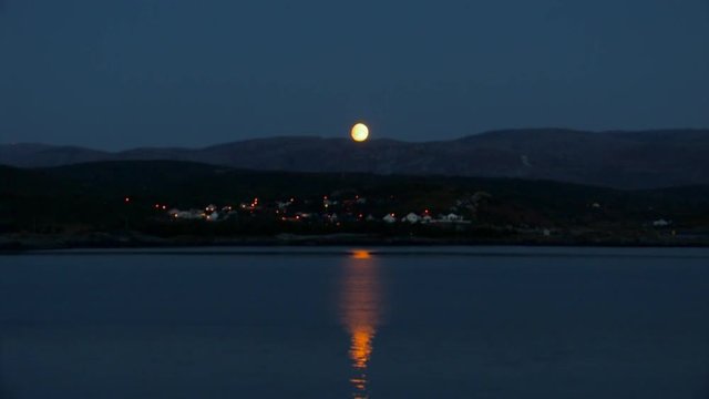 WIDE VIEW of a sleepy village on the coast. The moon glows and is reflected on the surface of the water as it sets behind a distant mountain range.