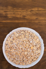 Oat flakes poured into a white porcelain bowl on a wooden background, top view.