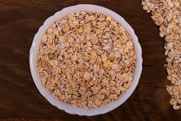 Oat flakes closeup in white porcelain bowl on wooden background, top view.
