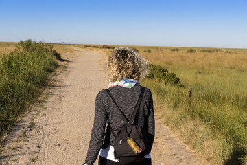 Frau wandert mit einem Rucksack einen sandigen Weg zwischen Salzwiesen an den Strand der Norsaee