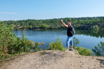 Fototapeta na wymiar Attractive senior white man with backpack stands on the rock and making selfie at the lake