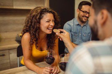 Beautiful girl with curly hair is smiling while her friend is feeding her with cracker. Friends having fun with red wine.