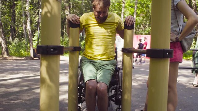 Young disable man in the park with his wife. The man is doing exercises on the parallel bars, the woman helping him.