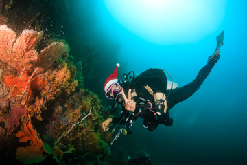 A female SCUBA diver wearing a santa hat for Christmas swimming over a colorful tropical coral reef