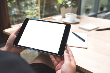 Mockup image of a woman's hands holding black tablet pc with white blank screen with notebook and coffee cup on table