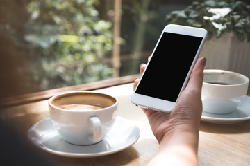 Mockup image of hand holding white mobile phone with blank black desktop screen with coffee cup on wooden table in cafe