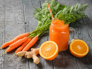 A mug of fresh carrot smoothie with cocktail straw, parsley, carrots, ginger root and oranges on a table.