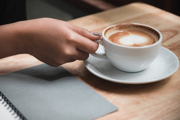 Closeup image of a woman's hand writing down on a white blank notebook while drinking coffee on wooden table