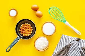 Ingredients for belgian waffles. Eggs, flour, sugar near waffle on pan, whisk for whipping on yellow background top view