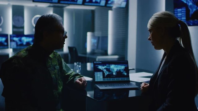 Female Special Agent Talks To Military Man in the Monitoring Room. In the Background Busy System Control Center with Monitors Showing Data Flow.