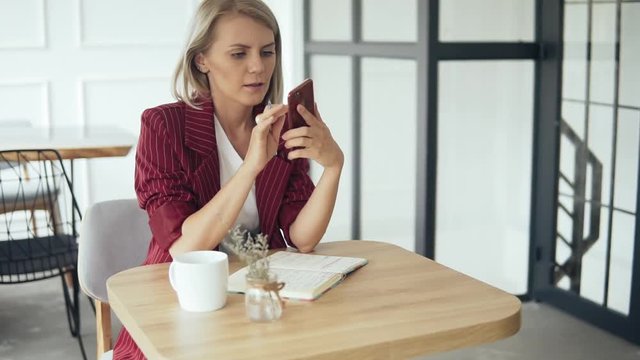 Beautiful and young successful business woman with mobile phone and notepad in a cafe, working as a freelancer. Blonde Female in red formal suit sitting at the table and using mobile application
