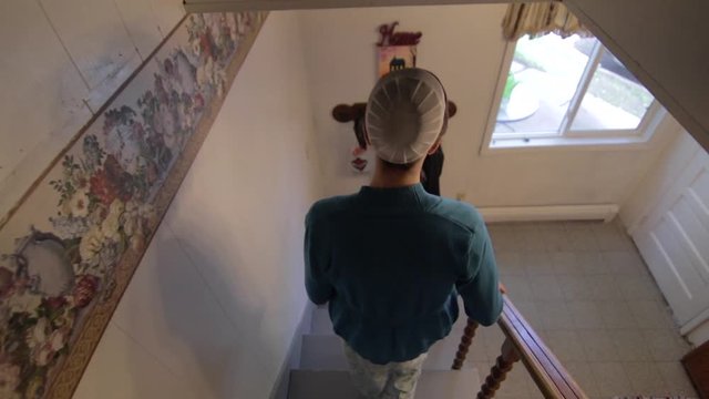 A young Mennonite woman walks down the stairs into a kitchen in slow motion.