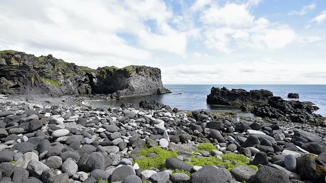 Landscape trail hiking view of rocky beach in Hellnar, National park Snaefellsnes Peninsula, Iceland with ocean sea waves, green grass in sunny summer day, cliff, clouds