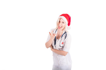 A young female doctor is isolated on a white background with a Santa Claus hat on her head. Medical concept of congratulations on Christmas and New Year. Hands show gesture of goat, tongue shows