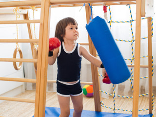 A small child is engaged in boxing on a wooden home sports complex. Girl in boxing gloves hits the...