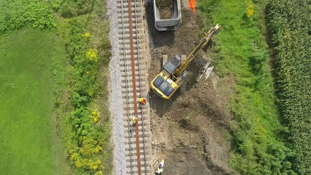Aerial: Flying over excavator working at railway reconstruction site. Filming rails renovation at railway beside beautiful river on sunny day.