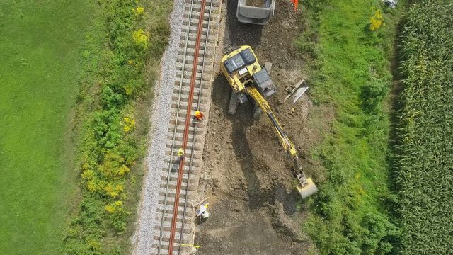 Aerial: Flying over excavator working at railway reconstruction site. Filming rails renovation at railway beside beautiful river on sunny day.