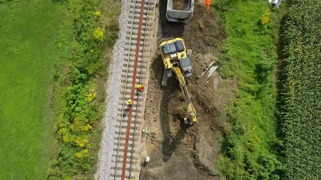 Aerial: Flying over excavator working at railway reconstruction site. Filming rails renovation at railway beside beautiful river on sunny day.